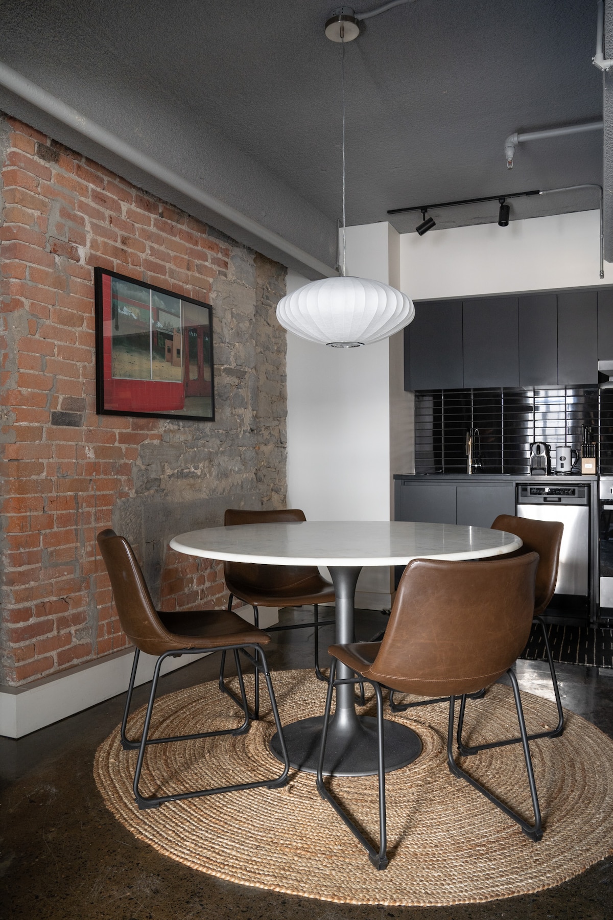 A dining area is highlighted by a round table surrounded by four brown chairs, situated on a woven rug. The space features exposed brick walls and modern kitchen cabinetry in the background, with a pendant light providing illumination.