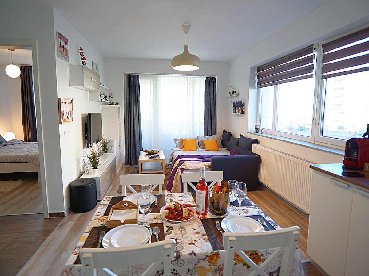 A modern living space features a cozy seating area with a dark blue sofa adorned with yellow and purple cushions. Natural light fills the room through large windows, and a dining table is set with plates and glasses, creating an inviting atmosphere for meals.