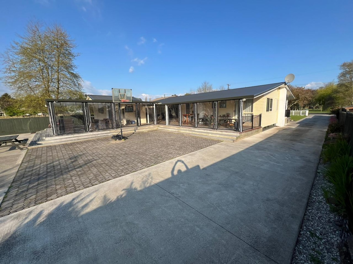 The exterior of the accommodation features a spacious cobblestone area, ideal for outdoor activities. A basketball hoop stands on one side, while the covered deck offers ample seating. The property is surrounded by greenery, providing a peaceful setting in the country.