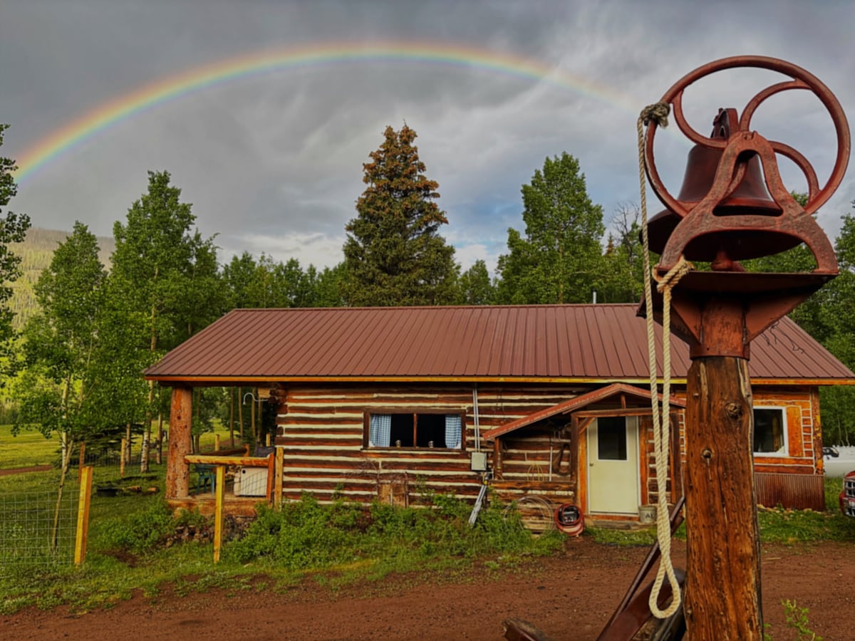 A cabin made of logs is framed by lush greenery and features a metal roof. A vintage bell is positioned in the foreground, with a rainbow arching across a cloudy sky above. The tranquil setting reflects the quiet surroundings of the property.