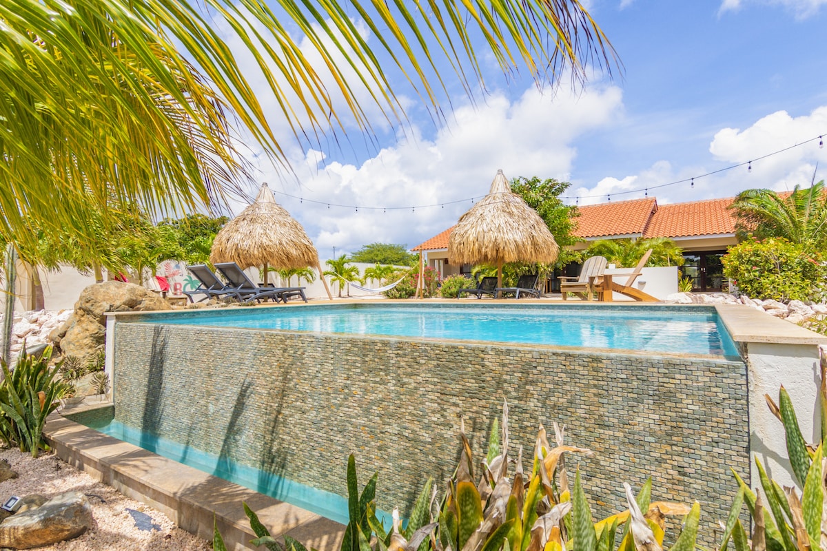 An infinity pool is showcased, surrounded by tropical plants and palm trees. Two thatched-roof cabanas are visible, offering shaded seating. The pool features a stone border, with clear water reflecting the sky and the vibrant greenery around.