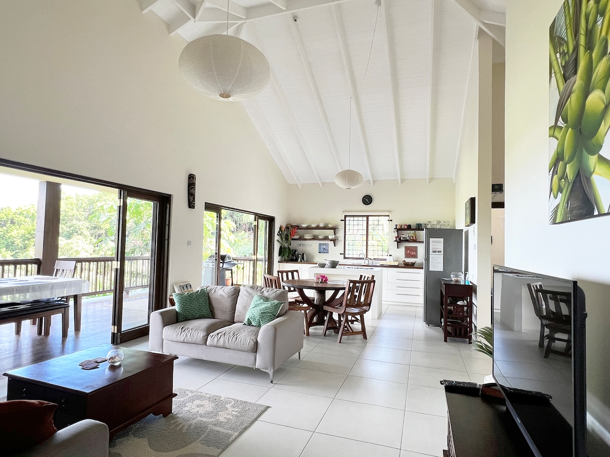 An open living space features high vaulted ceilings and ample natural light. A cozy seating area is arranged with a gray sofa and a coffee table, adjacent to a dining area with a wooden table and chairs. Large glass doors lead to a scenic outdoor deck.