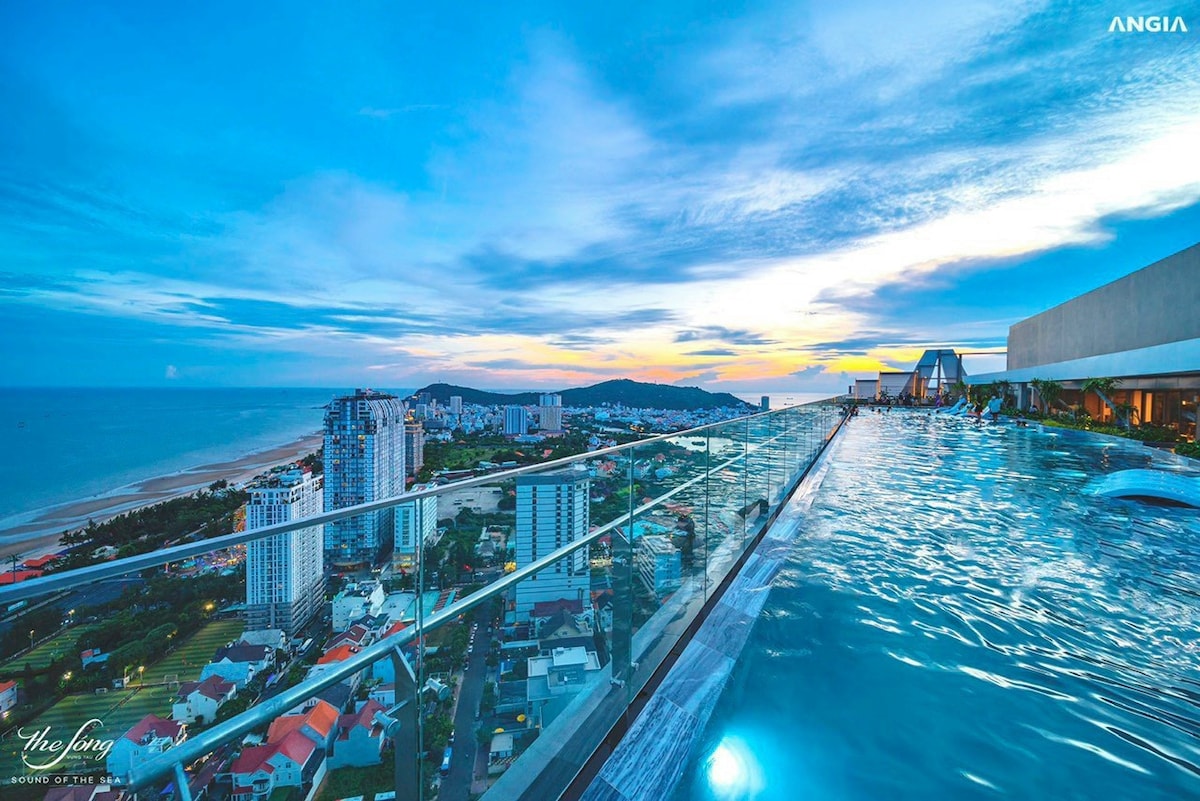 An infinity swimming pool is visible on the rooftop, offering expansive views of the coastline and skyline. The water reflects the vibrant colors of the sunset, while glass railings ensure safety without obstructing the scenic landscape.
