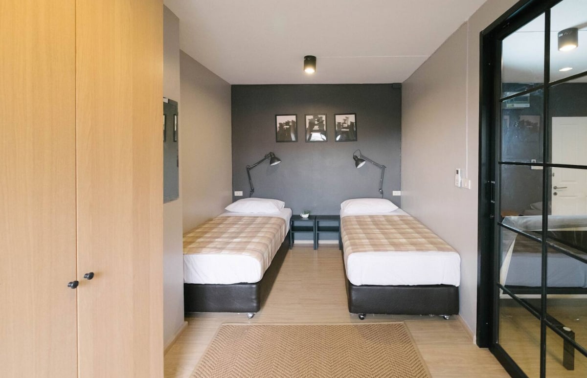 A private room is showcased with two twin beds, each featuring a neutral-toned plaid blanket. A small table is positioned between the beds, while two adjustable reading lamps stand on either side. A large wardrobe is visible on the left, and framed photographs decorate the wall behind.