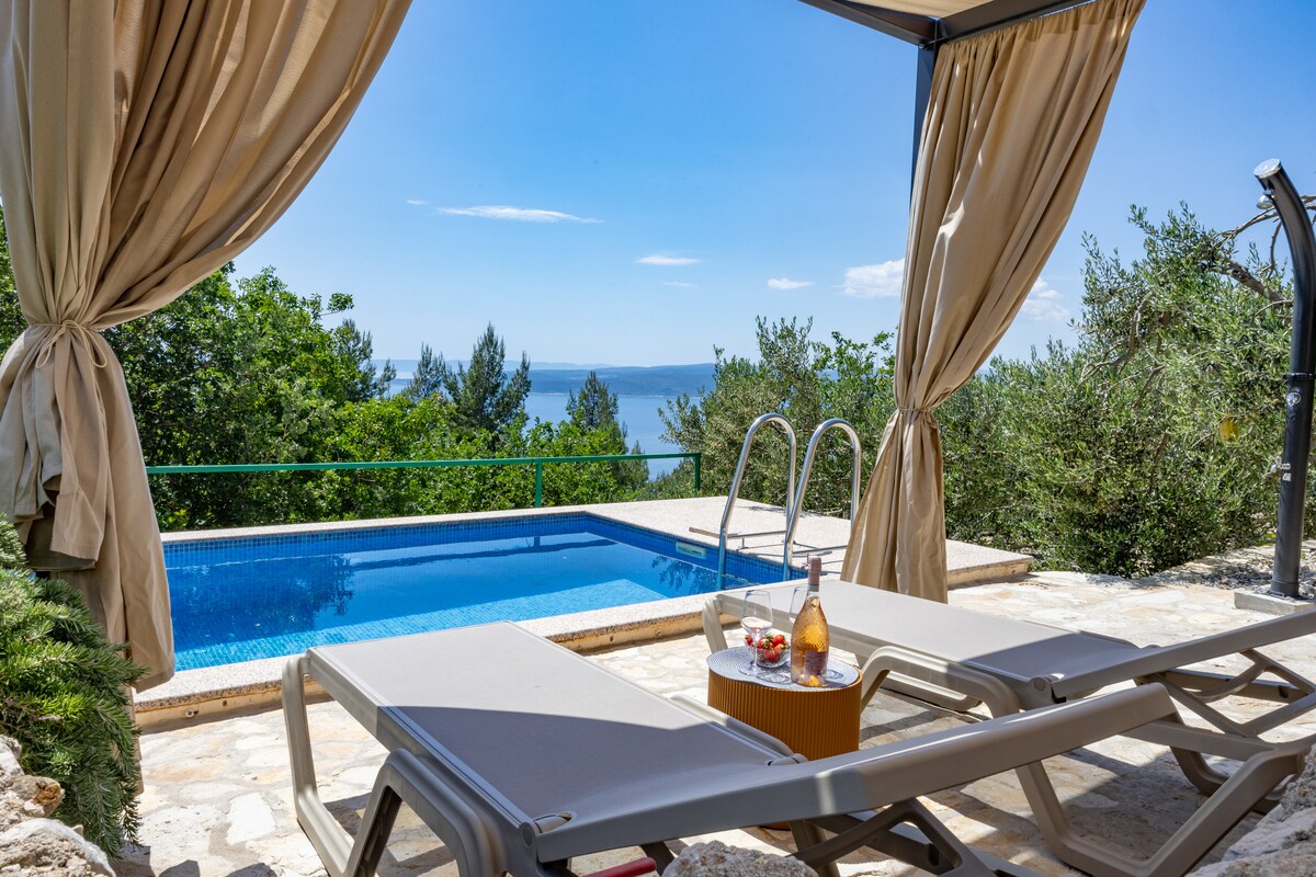 A private terrace features sunbeds and shade, overlooking a sparkling swimming pool. Lush greenery surrounds the area, providing a serene backdrop, while the sea is visible in the distance. A small table holds refreshments, enhancing the outdoor experience.