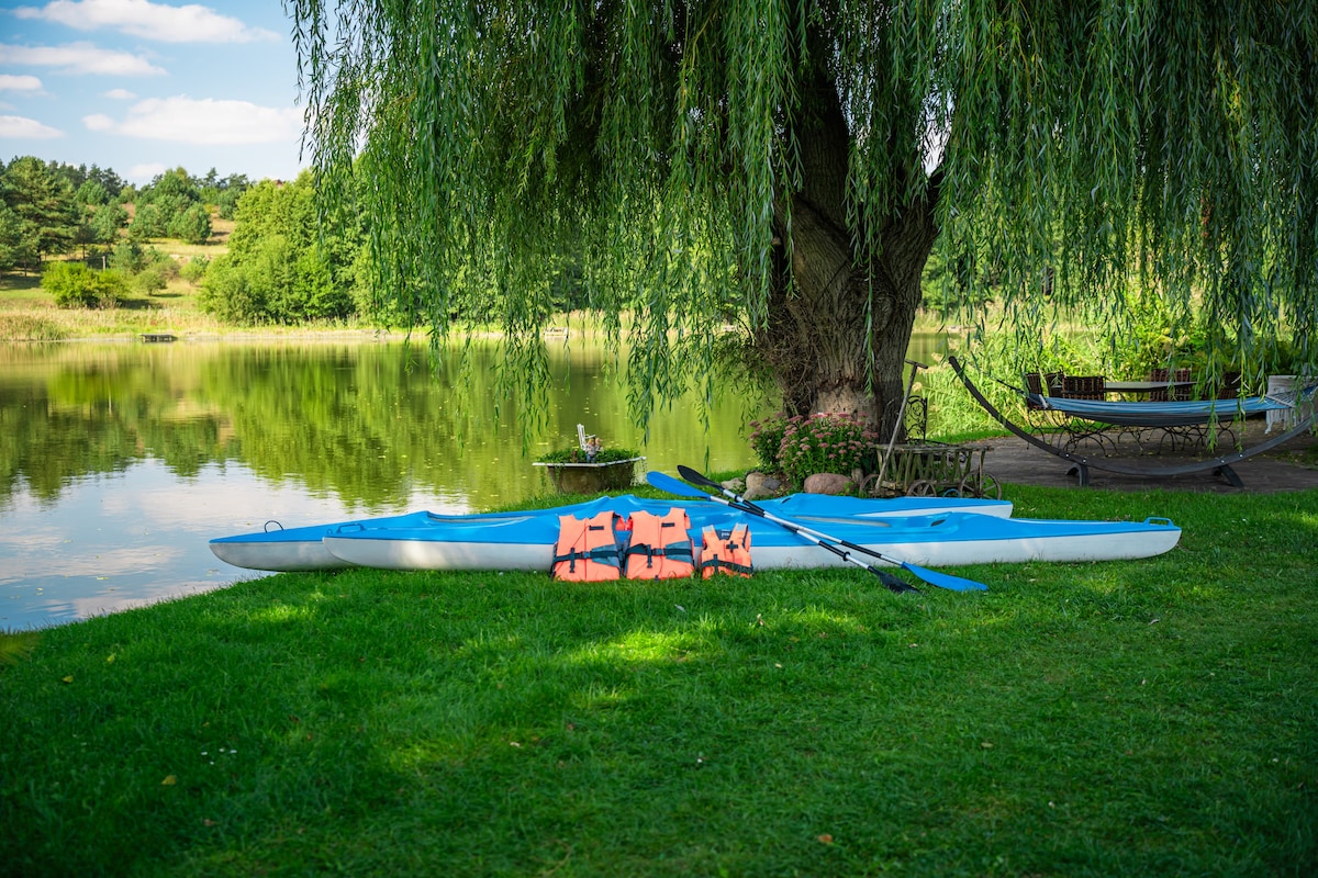 Two kayaks, one blue and one white, are positioned on verdant grass near a tranquil lake. Bright orange life vests are neatly arranged on the kayaks. A large willow tree provides shade, and a glimpse of the water reflects the surrounding greenery.