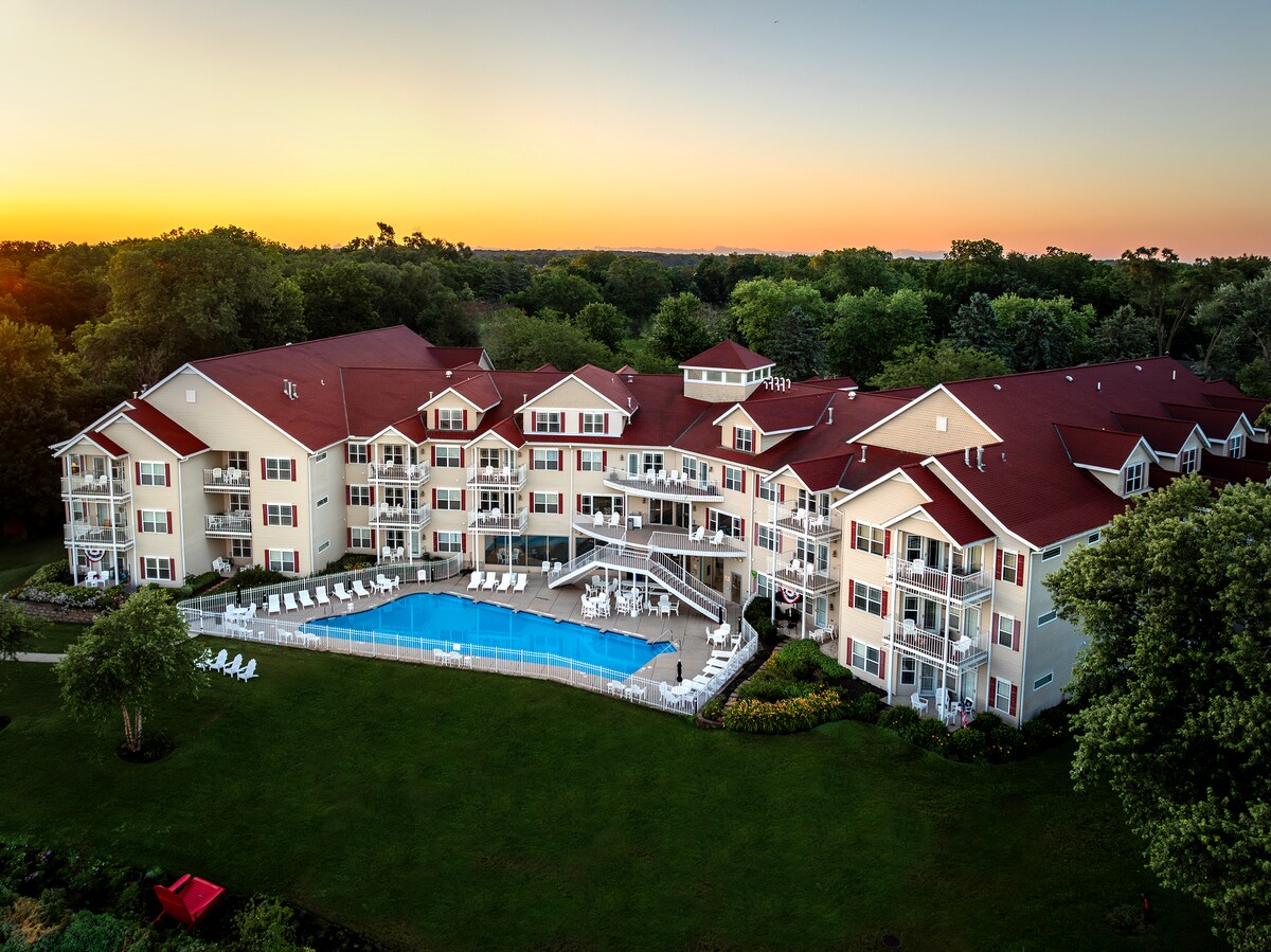 The resort features a multi-story building with a red roof and white walls, surrounded by lush greenery. An outdoor pool is visible in the foreground, with lounge chairs arranged around it. The setting offers a serene atmosphere, highlighted by a colorful sunset in the background.