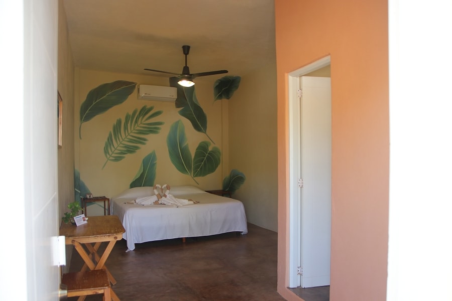 A spacious room is presented, featuring tropical leaf murals on the walls. A king bed dressed in white linens is positioned centrally, complemented by a ceiling fan above. A simple wooden table is placed nearby, and an entryway leads to additional areas within the accommodation.