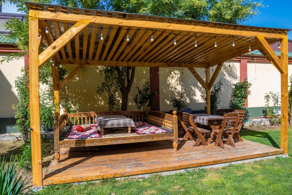 A wooden gazebo features a spacious lounging area with traditional patterned cushions and a low table. Soft lighting is provided by string lights overhead. Nearby, a dining table with wooden chairs is set on a well-maintained lawn, surrounded by lush greenery.