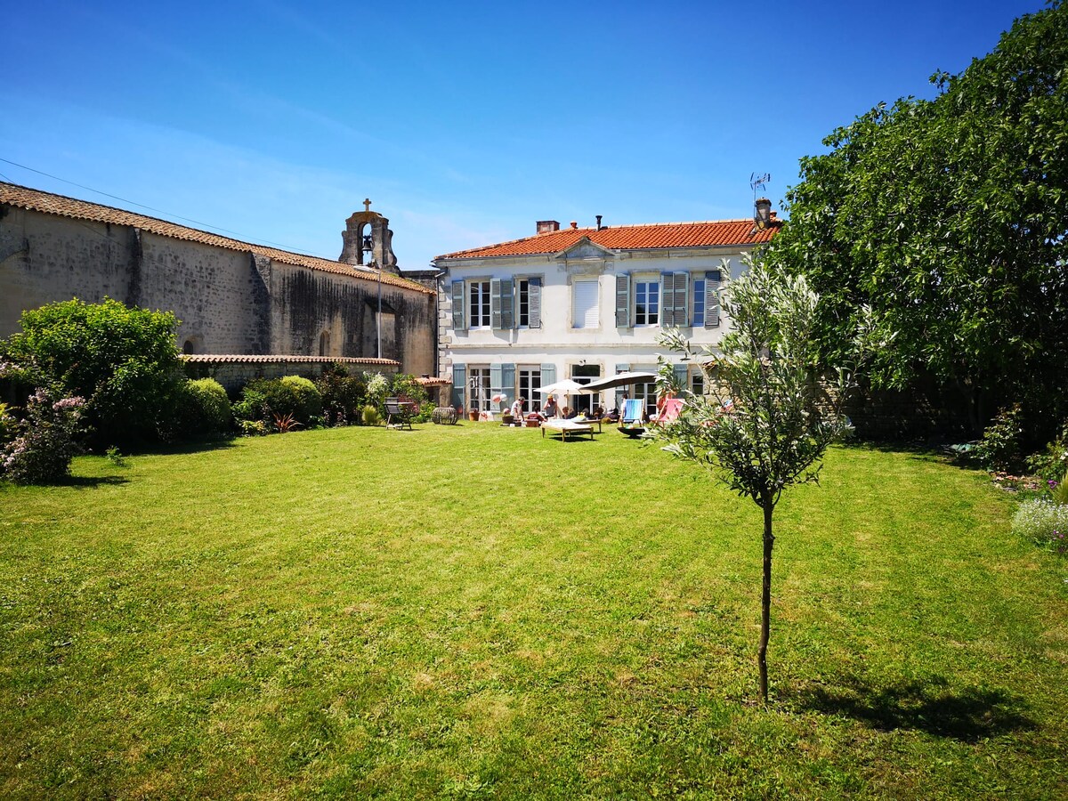 The exterior of the 18th-century Presbytère is captured, featuring a spacious garden adorned with lush greenery. The house, characterized by large windows, offers seating areas that overlook the garden. A tree stands at the forefront, enhancing the tranquil ambiance of the outdoor space.