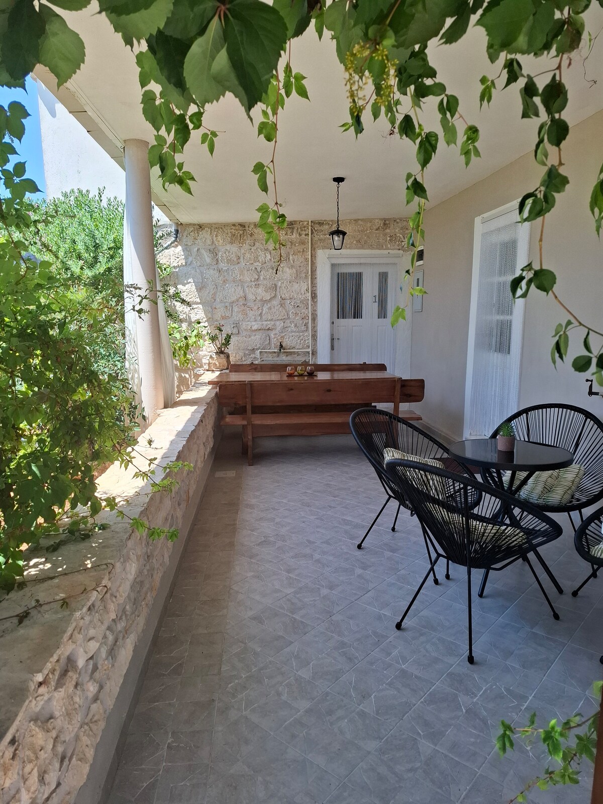 A covered terrace features a wooden bench and a round table surrounded by four black chairs. Lush greenery frames the space, providing a natural touch. Neutral tile flooring complements the light-colored walls, while a door leads to the interior of the apartment.
