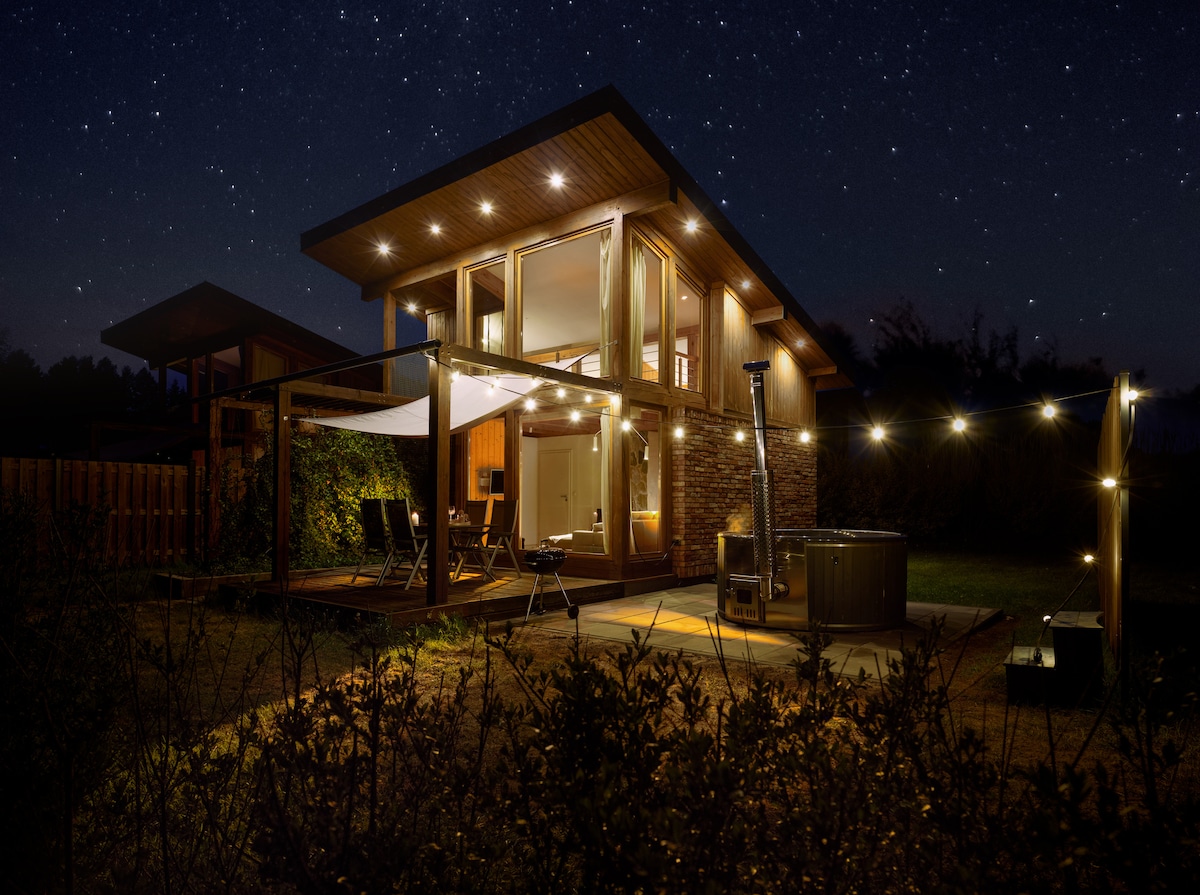 A modern wooden cabin is depicted at night, featuring warm lights illuminating the exterior. A spacious patio is highlighted, equipped with seating and a hot tub, surrounded by landscaped gardens. The starry sky adds a serene backdrop to this tranquil setting.