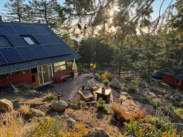 A serene outdoor space features a flagstone patio surrounded by boulders and native plants. Solar panels are installed on the roof, while sunbeams filter through the trees, creating a peaceful ambiance. Comfortable seating is arranged for relaxation, with nature views in the background.