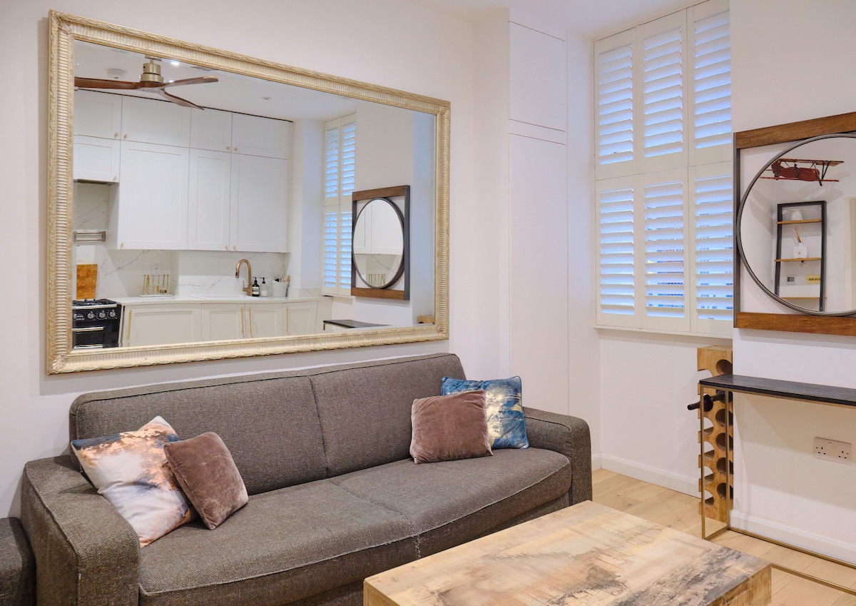 A spacious living area features a comfortable sofa adorned with decorative pillows. A large mirror enhances the light in the room, while a coffee table made of natural wood occupies the center. Light-colored cabinetry is visible in the kitchen area, enhancing the overall brightness.