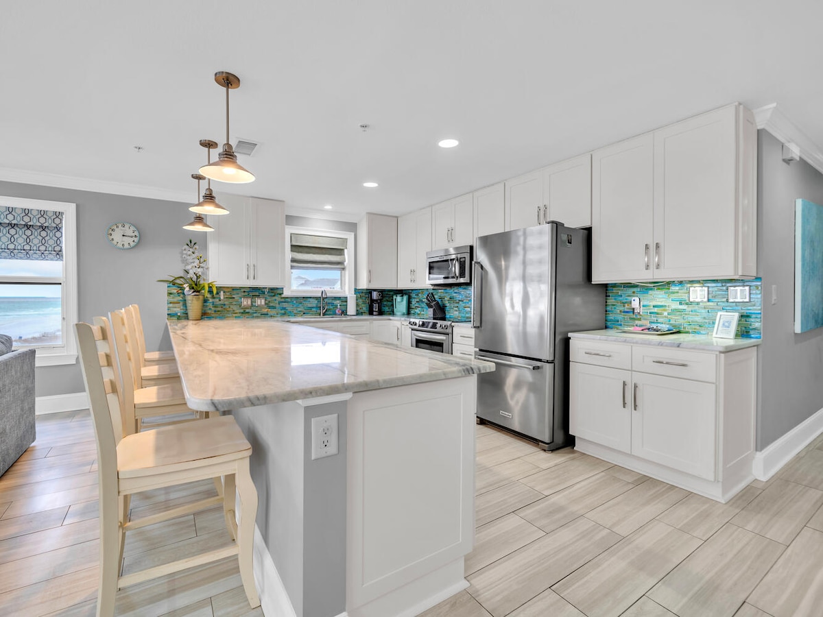 The kitchen features modern white cabinetry and a spacious island with seating. Stainless steel appliances are arranged thoughtfully, and colorful backsplash tiles add a touch of vibrancy. Natural light illuminates the space, highlighting the contemporary design and functional layout.