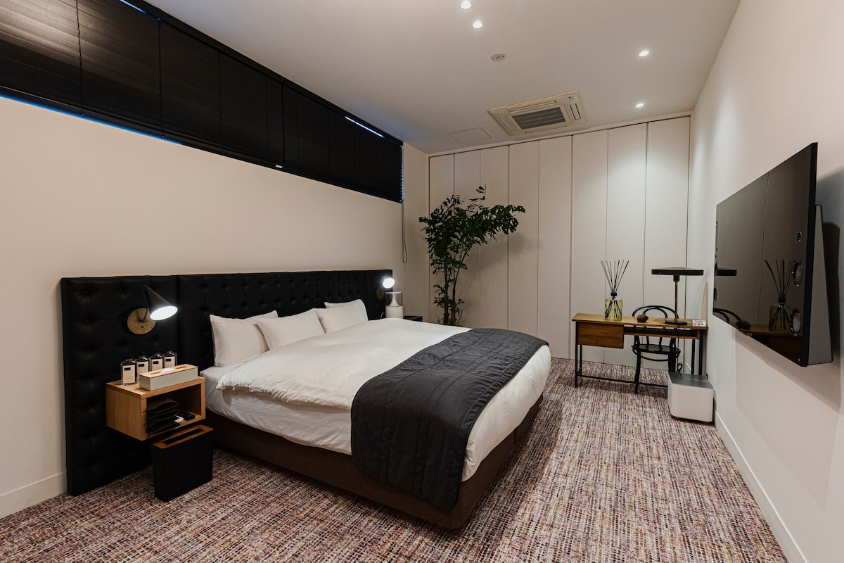 A spacious sleeping area features a large bed with a dark headboard and neutral bedding. A wall-mounted television is positioned opposite the bed. Natural light filters through black blinds, and a small plant adds a touch of greenery to the room.