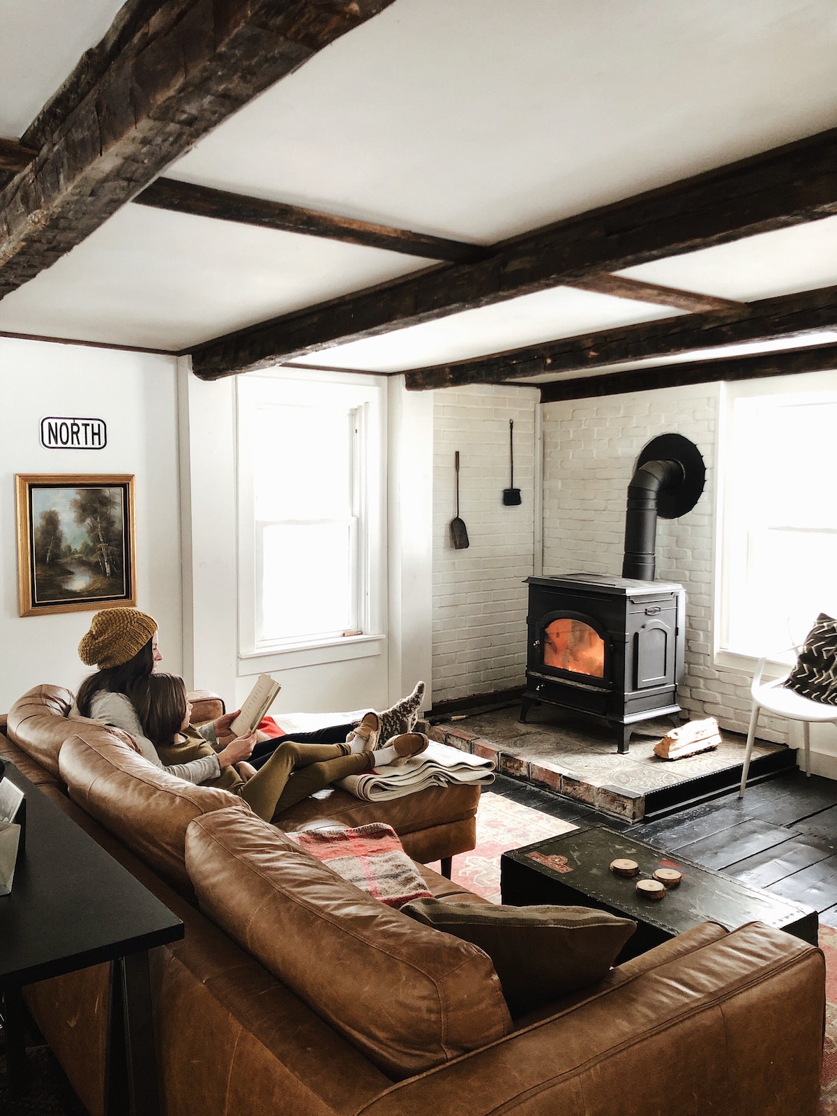 A cozy living room features original exposed beams and a wood-burning stove. A large leather sofa is complemented by a patterned rug, while natural light fills the space through the windows. A vintage artwork hangs on the wall, adding character to the room.