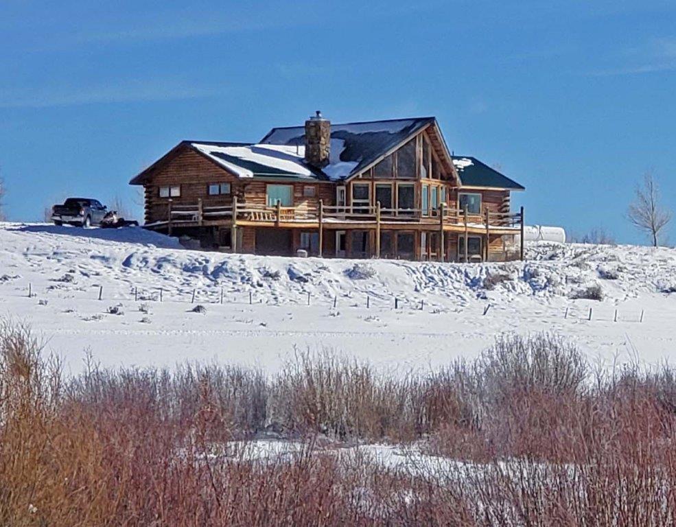 A spacious lodge is set against a backdrop of snow-covered ground and clear blue skies. The building features a combination of stone and wood materials, with multiple balconies and large windows that invite light. Surrounding vegetation includes low shrubs and open landscape.