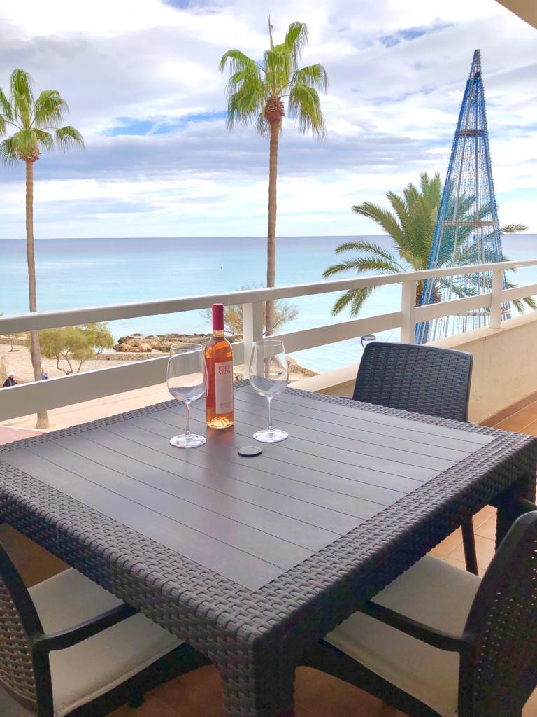 A waterfront balcony features a table set for two, complete with glasses and a bottle of wine. Palm trees frame the view, while the serene sea and sky create a calming backdrop. The outdoor dining area invites relaxation and enjoyment of the surroundings.