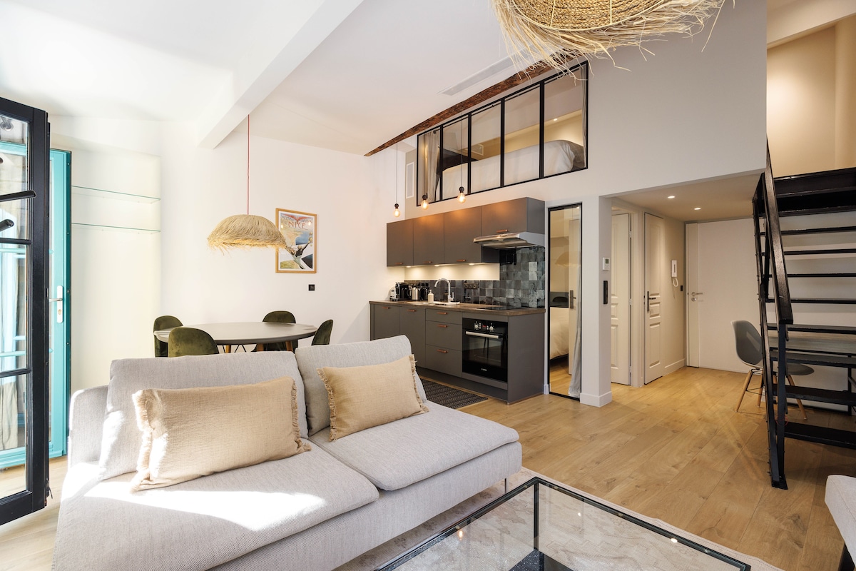 An open-plan living area features a comfortable sofa with decorative cushions and a glass coffee table. The kitchen area includes modern appliances, complemented by a dining space with a round table and green chairs. A staircase leads to an upper sleeping area, enhancing the apartment's spacious feel.