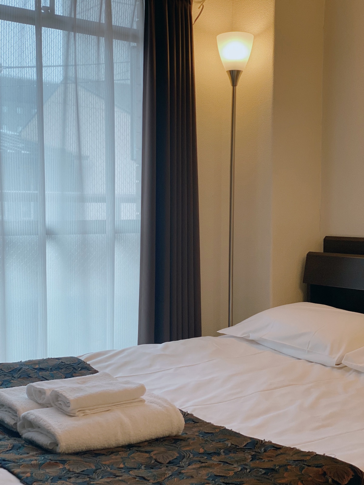 A comfortable sleeping area features a single-sized bed, adorned with fresh linens. A bedside lamp provides soft lighting, and sheer curtains allow natural light to filter in. A folded towel and a decorative cover are neatly arranged on the bed.