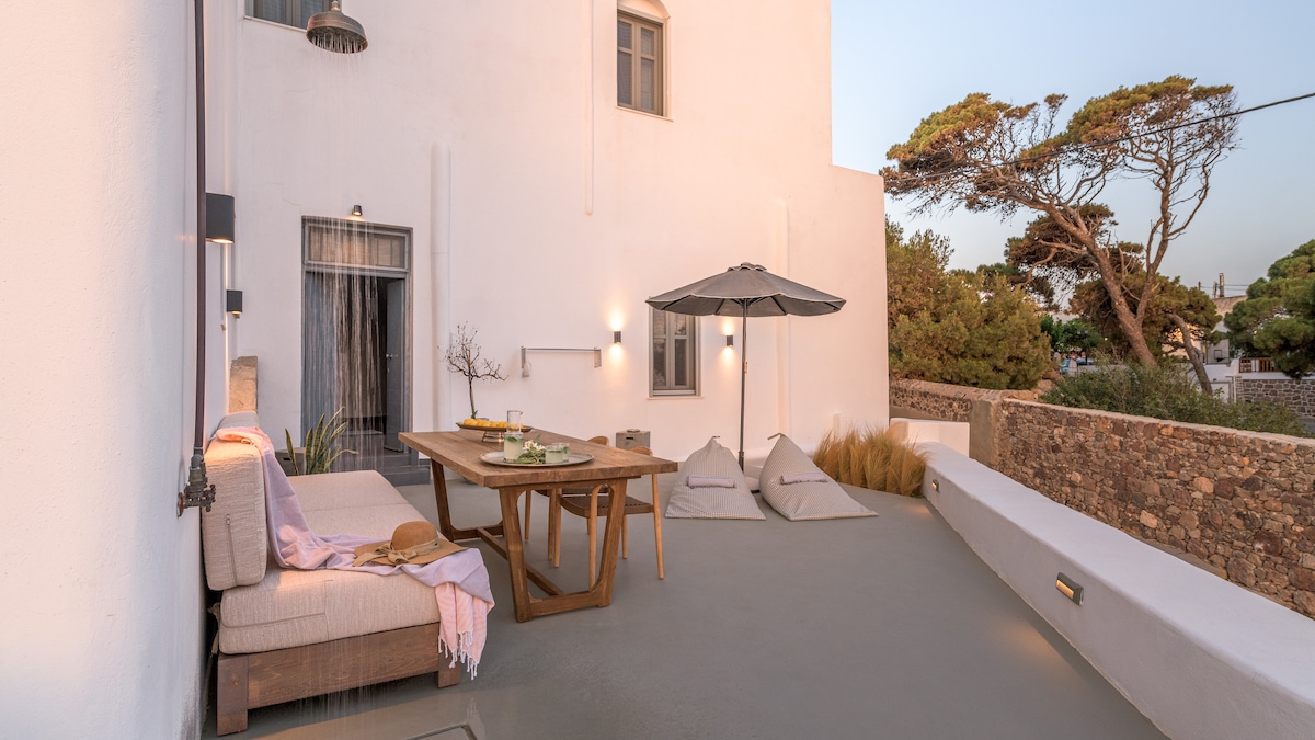 A private outdoor area features a wooden dining table surrounded by comfortable seating cushions. An umbrella provides shade, while a soft throw is draped over the corner couch. The serene setting is complemented by rustic stone walls and greenery in the background.