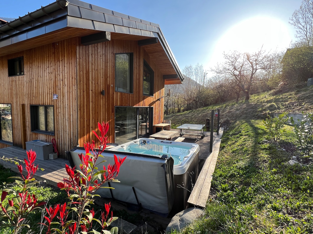 The exterior of the chalet features wooden paneling and large glass windows. A jacuzzi is positioned on a wooden deck, surrounded by greenery and vibrant flowers. The sun casts a warm glow across the space, creating a peaceful outdoor setting.
