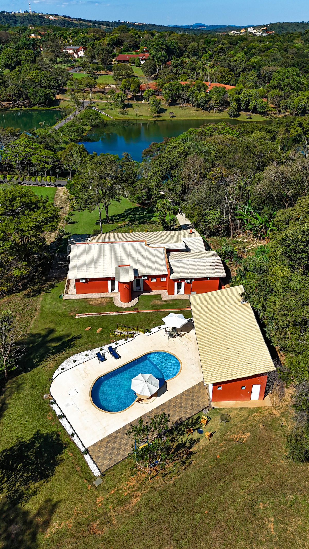 An aerial view reveals a spacious, red-painted house with a large pool surrounded by tiled decking. Lush greenery and lakes are visible in the background, enhancing the tranquil setting of the property.