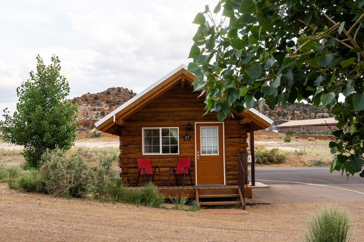 A charming cabin with a wooden exterior is framed by green trees and shrubs. Two red chairs sit on the porch, overlooking a gravel path. A bright orange door and multiple windows allow natural light to fill the interior. Scenic rock formations are visible in the background.