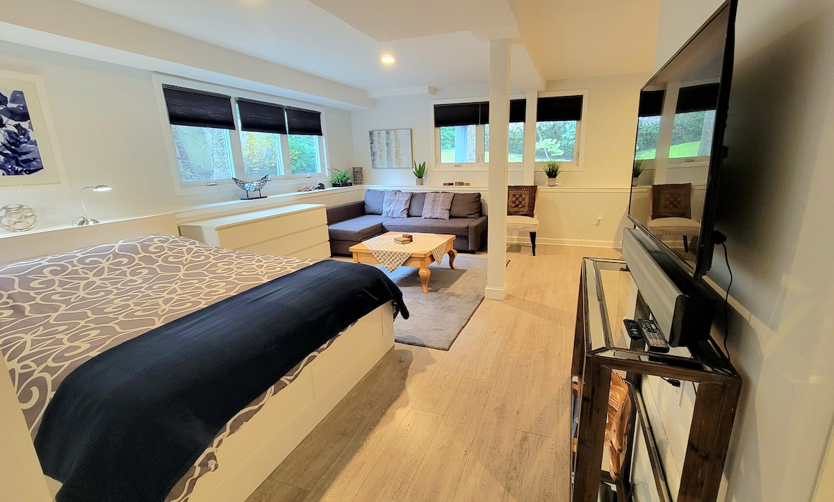 A spacious guest suite is shown, featuring a cozy sleeping area with a large bed covered in neutral linens. A comfortable seating area with a sectional sofa and a wooden coffee table is visible, alongside a TV mounted on a stand. Natural light enters through large windows.