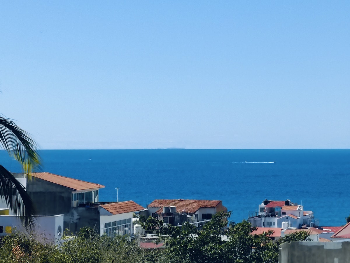 A wide view of the ocean is presented, featuring a clear blue sky and gentle waves. The shoreline is dotted with rooftops from nearby buildings, showcasing the coastal landscape. Some lush greenery is visible in the foreground, complementing the serene atmosphere.