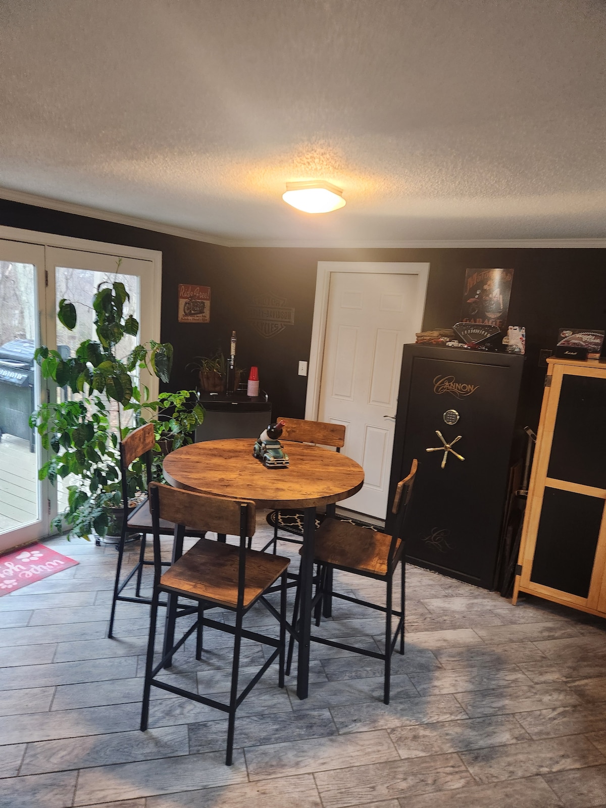 A dining area features a round wooden table surrounded by four tall chairs. Natural light enters through large glass doors, illuminating the black walls. A vibrant green plant stands in the corner, and a dark cabinet is visible, adding functional storage to the space.