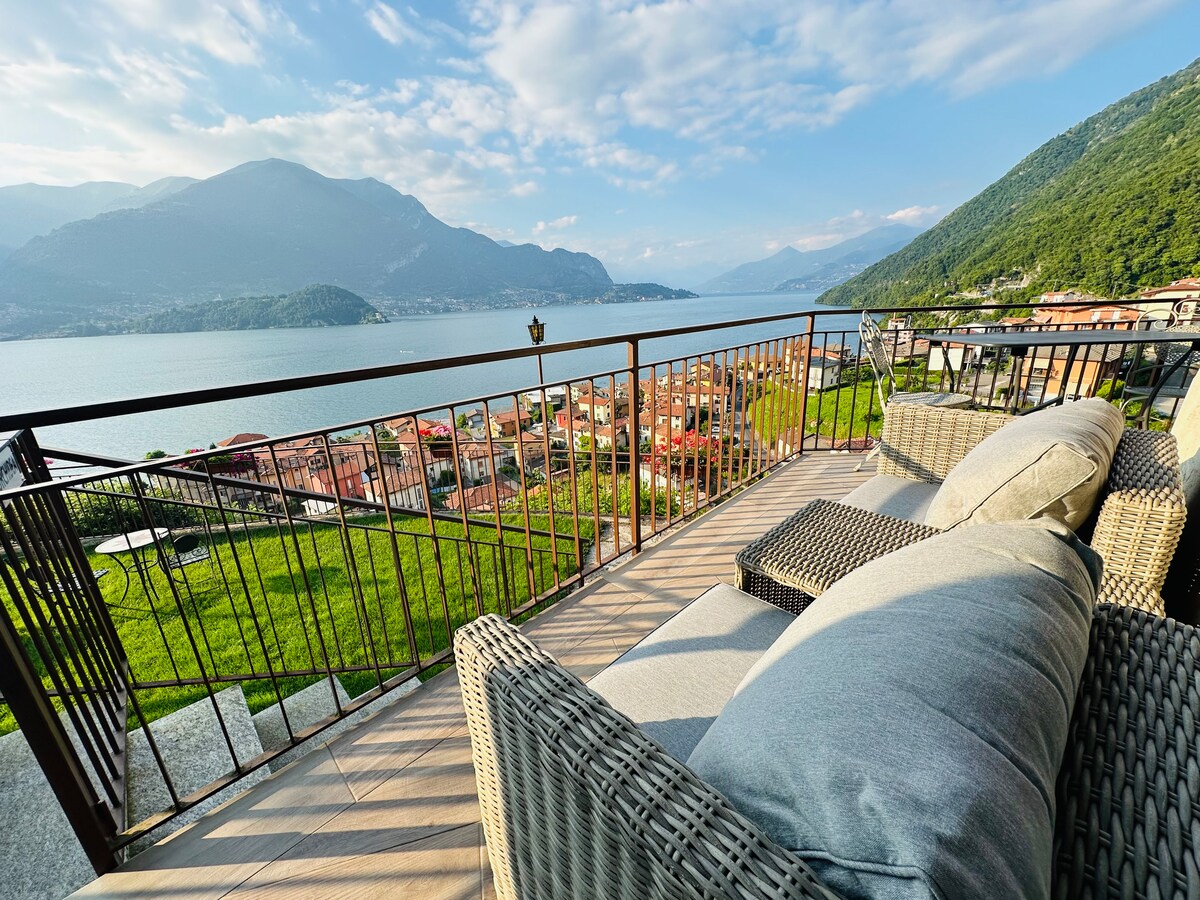 A private terrace is furnished with a cozy seating area, offering a stunning view of the lake and surrounding mountains. The railing frames the picturesque landscape, while the soft cushions invite relaxation in this serene outdoor space.