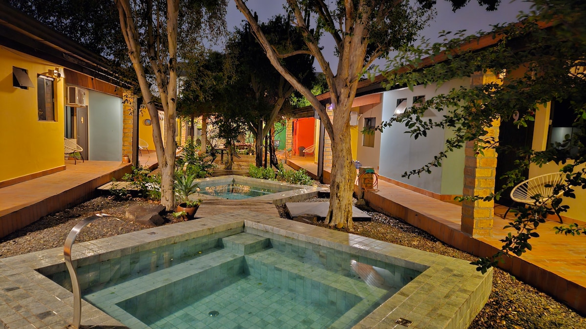 A serene courtyard is surrounded by small, brightly colored buildings. Water features can be seen gently flowing, with pathways leading to various seating areas under the shade of trees. The layout invites relaxation and encourages social interaction among guests.