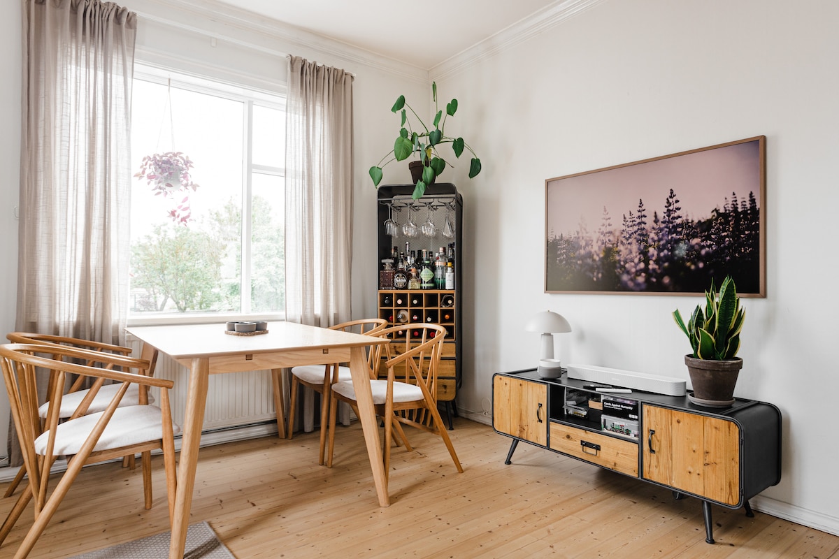 A bright dining area features a wooden table surrounded by three light chairs. A large window with sheer curtains allows for natural light, while a greenery accent sits on the window sill. A stylish entertainment unit displays decorative items and a large artwork.