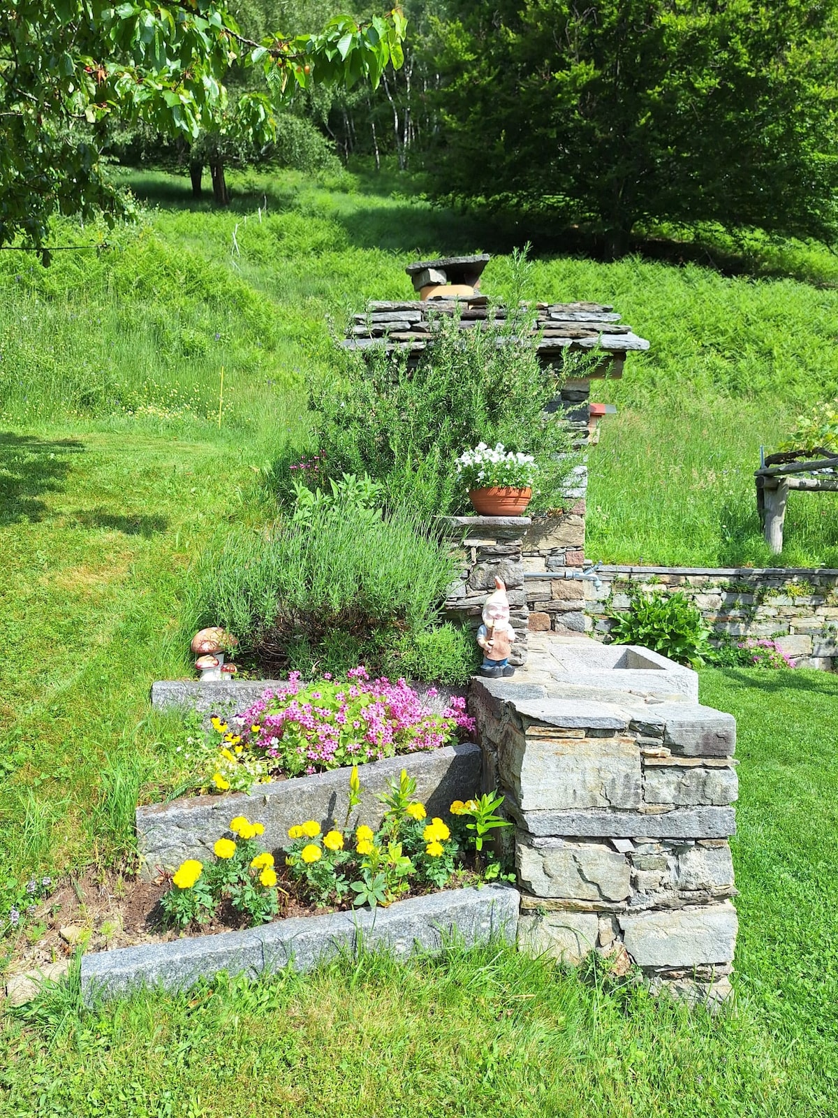 A stone garden features meticulously arranged flower beds with vibrant blooms, including yellow marigolds and purple petunias. Greenery surrounds the space, complemented by decorative pots and a rustic structure that nurtures herbs. The scene is set against the backdrop of lush, sloping meadows.