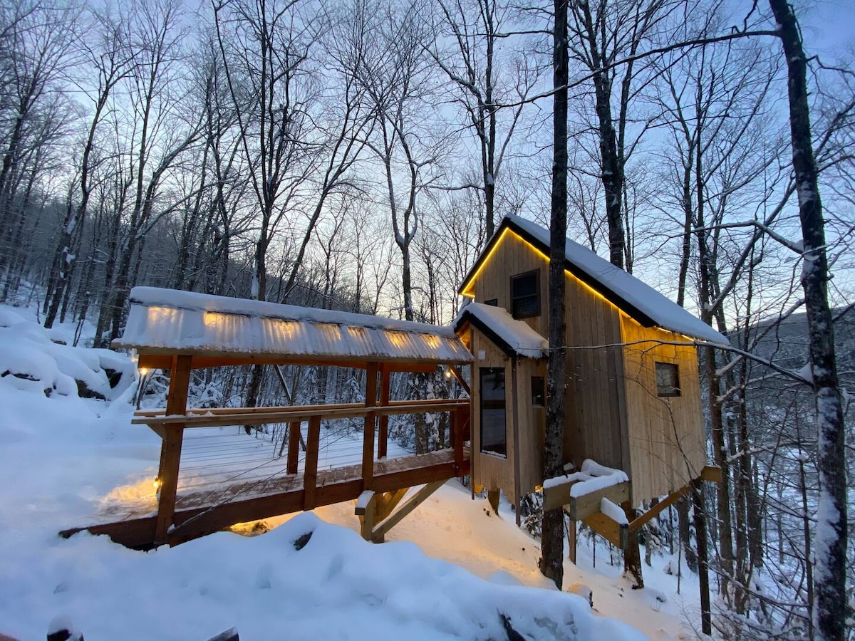 A cozy treehouse is nestled among bare winter trees, with a snow-covered pathway leading to its entrance. Soft light emanates from the windows, contrasting with the surrounding white landscape, creating a serene winter setting.