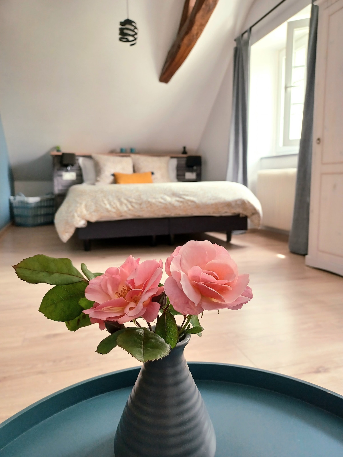 A spacious bedroom reveals a comfortable bed adorned with soft linens, complemented by decorative pillows. Natural light enters through the window, enhancing the room's calm appearance. A vase with pink flowers is placed on a blue tray in the foreground, adding a touch of color.