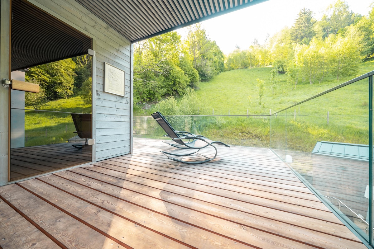 A spacious outdoor terrace features wooden flooring and a glass railing that provides a clear view of the lush green hillside. A reclining lounge chair is positioned for relaxation, offering a serene outdoor space that invites enjoyment of nature.
