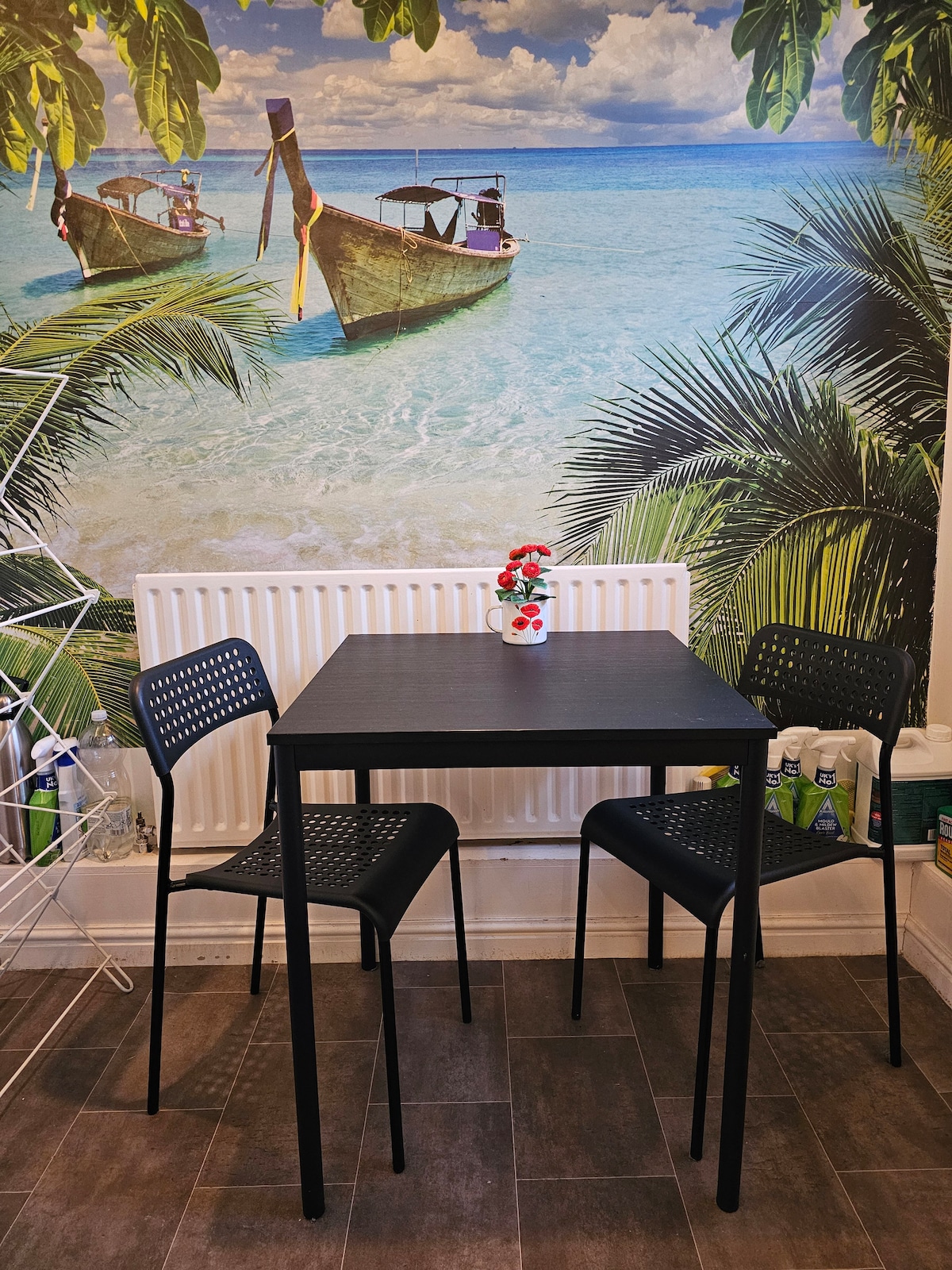A small dining area is displayed with a black table and two matching chairs, positioned against a vibrant wallpaper featuring a tropical beach scene. A vase with flowers adorns the table, while cleaning supplies are neatly arranged nearby.