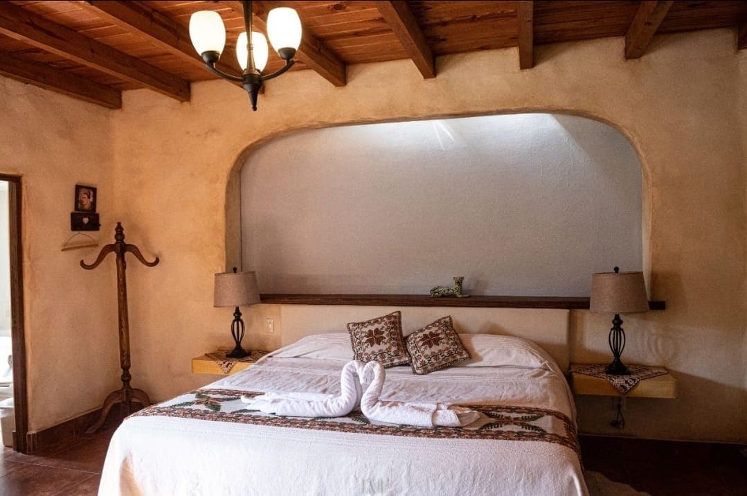 A cozy bedroom features a spacious bed adorned with a decorative blanket, while two bedside lamps offer soft lighting. Exposed wooden beams create a rustic ceiling. A curved alcove wall serves as a unique backdrop, complemented by an elegant wall-mounted shelf.
