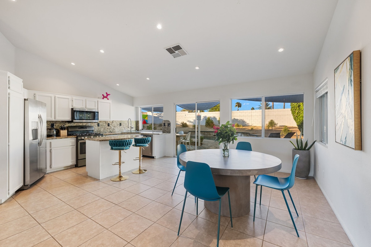 An open concept kitchen and dining area features a circular dining table surrounded by blue chairs. A large window illuminates the space, while a well-equipped kitchen is visible, highlighted by modern appliances and decorative tile backsplash. Potted plants add a touch of greenery.
