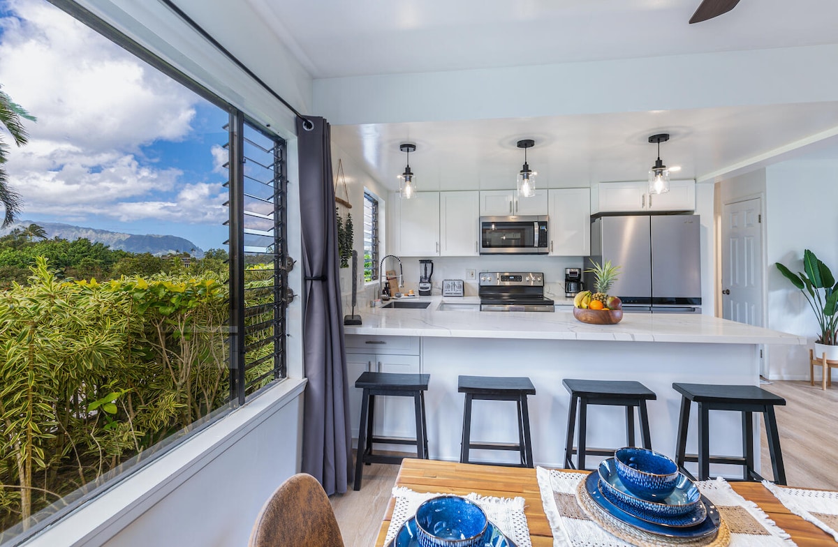 An open kitchen area features modern appliances and a spacious countertop, paired with a dining table set with blue dishware. Natural light floods the space through a large window, offering views of greenery and distant mountains.