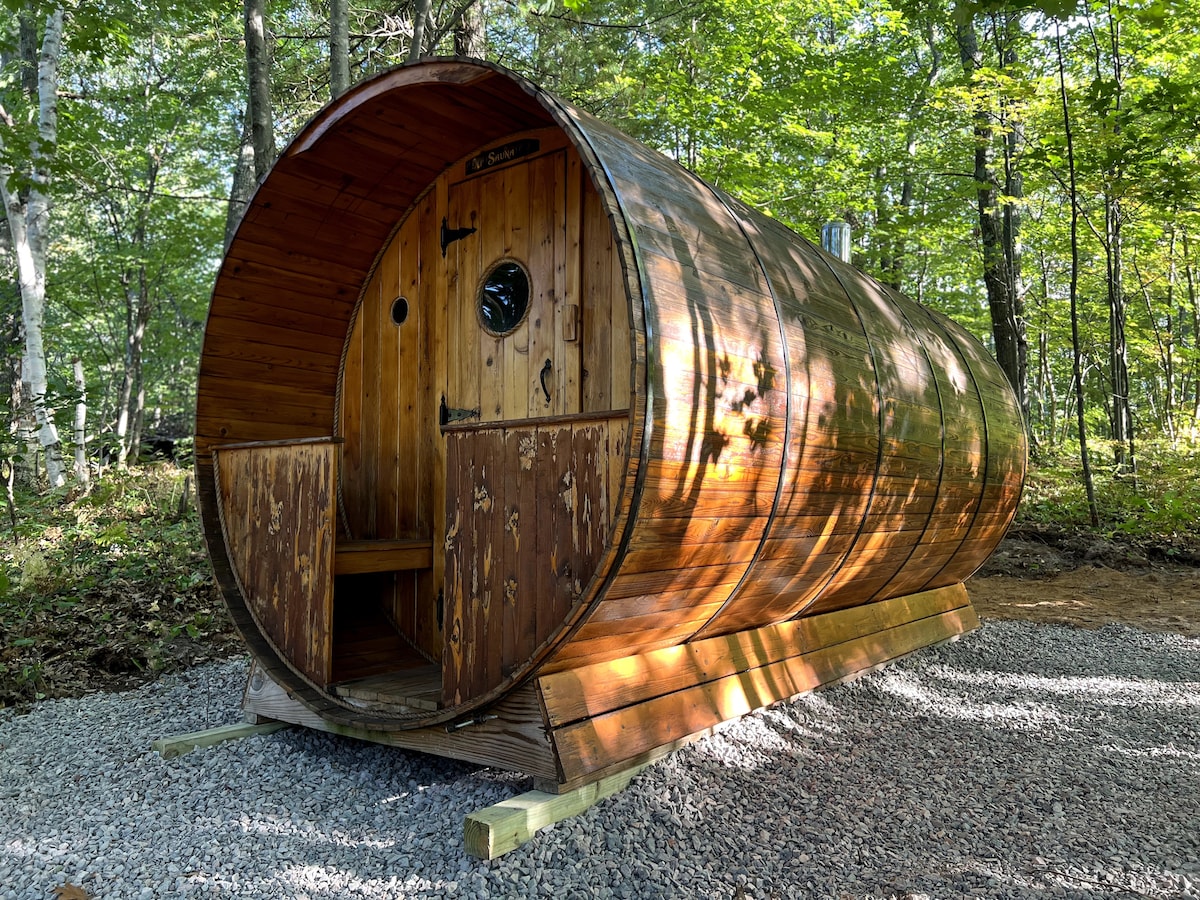 A barrel sauna constructed from wood is set on a gravel base amidst trees. The rounded design features a circular window and a wooden door, providing an inviting entrance. Natural wood grain is visible, showcasing the sauna's rustic charm.