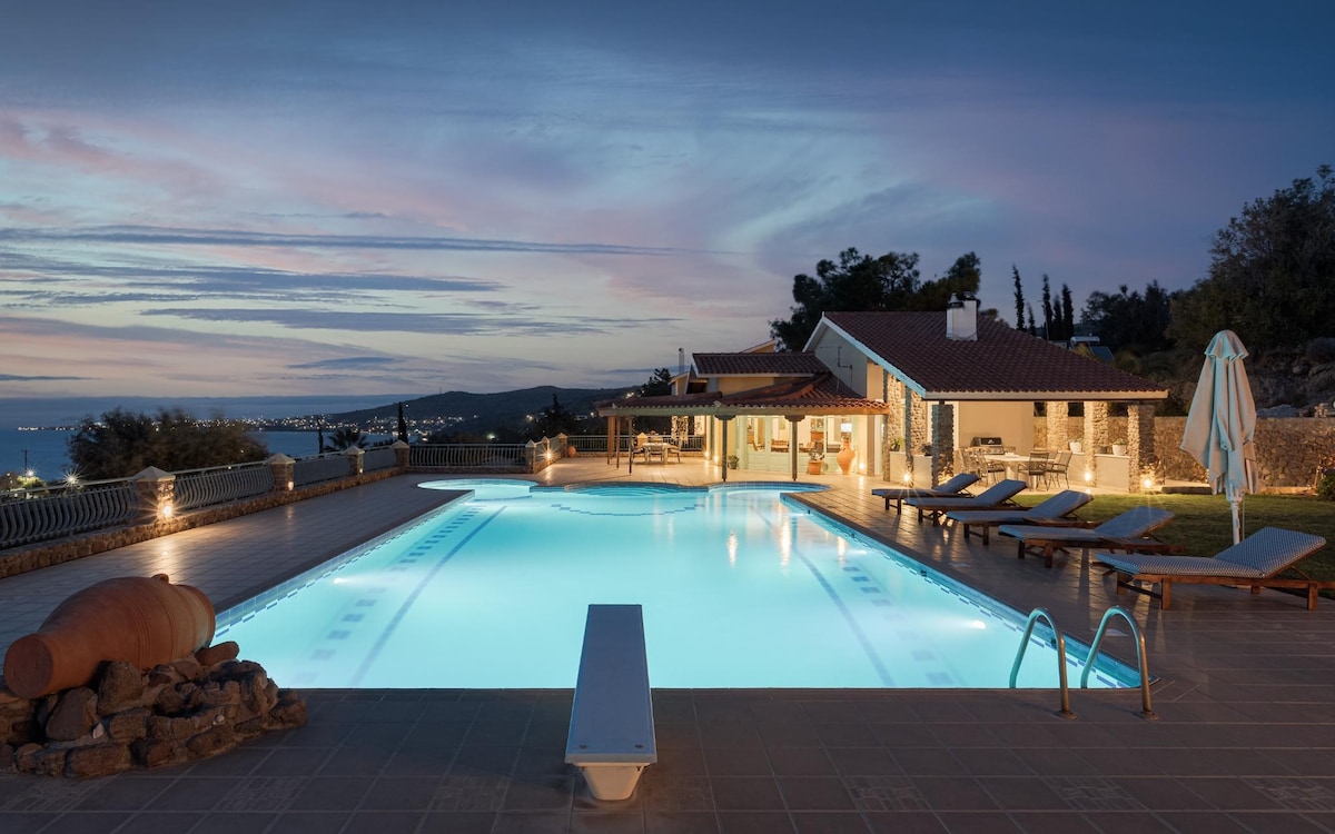 A large pool area is illuminated in soft blue tones against a dusky sky, featuring sleek lounge chairs lined along the tiled deck. A pool house with large windows connects to the serene outdoor space, while gentle landscape forms a backdrop.