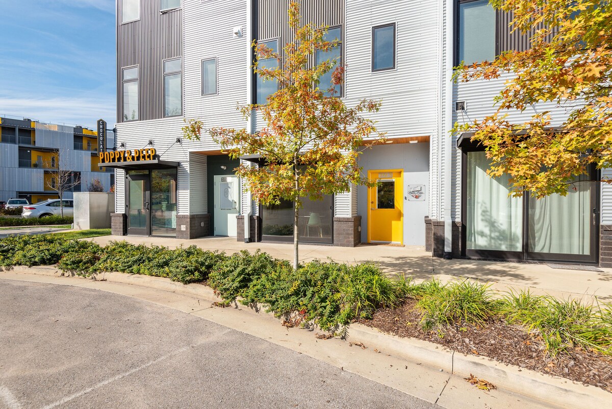 The exterior of Cocoa Casa is shown, featuring contemporary architecture with a mix of metal and glass. A vibrant yellow entrance is visible, framed by large windows and landscaping that includes shrubs and small trees. The surrounding area reflects a modern industrial aesthetic.