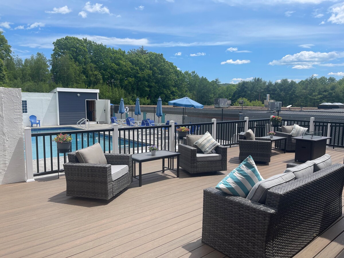 A spacious deck area features comfortable seating arrangements with dark wicker furniture and cushions. The setting overlooks a pool area, equipped with umbrella-covered lounge chairs. Lush greenery surrounds the space, providing a relaxing atmosphere under a clear blue sky.