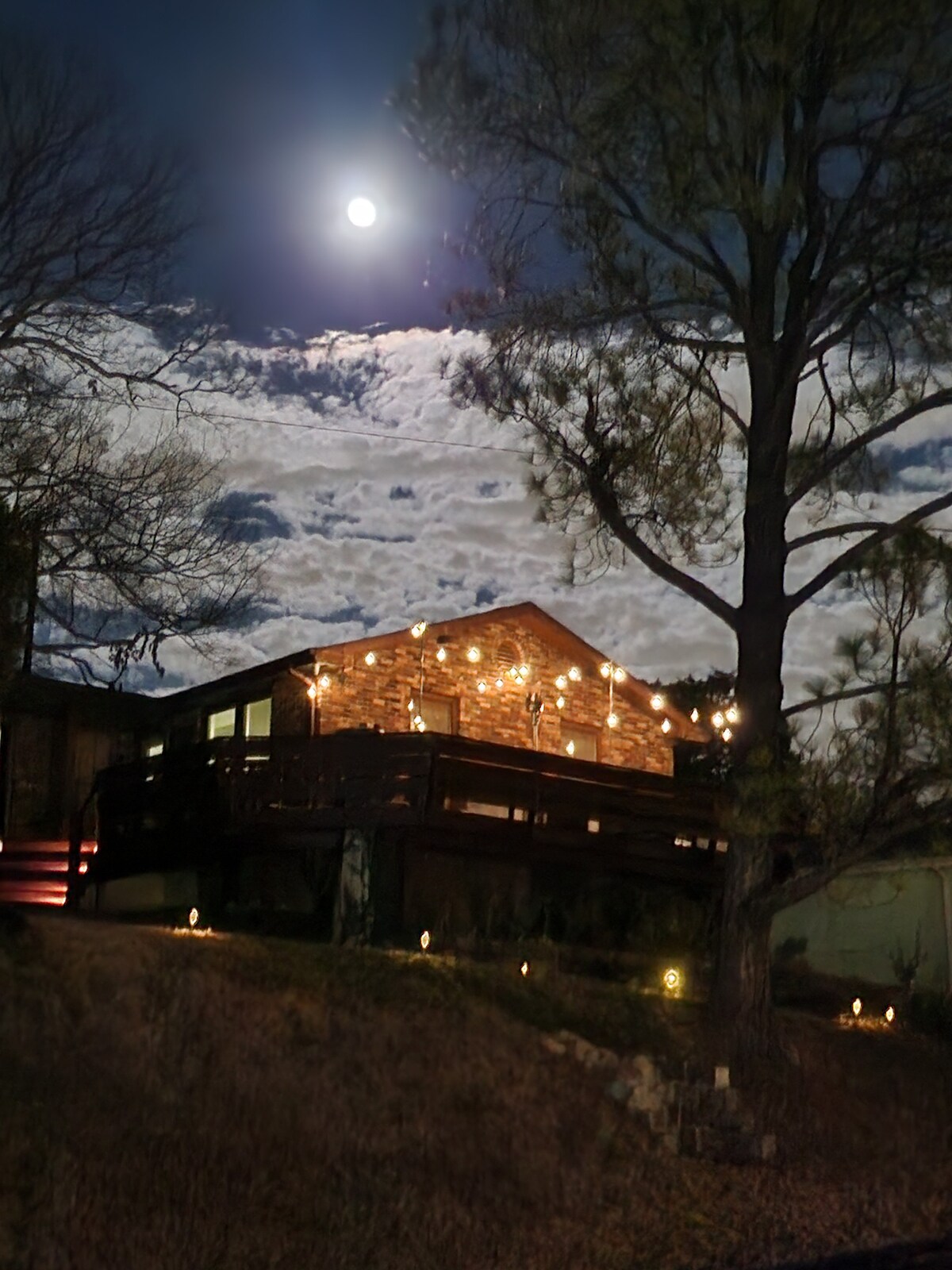 A charming house illuminated by soft outdoor string lights stands against a night sky. The full moon shines brightly above, casting light on the surrounding trees and clouds, while the structure features large windows that invite a warm glow from within.
