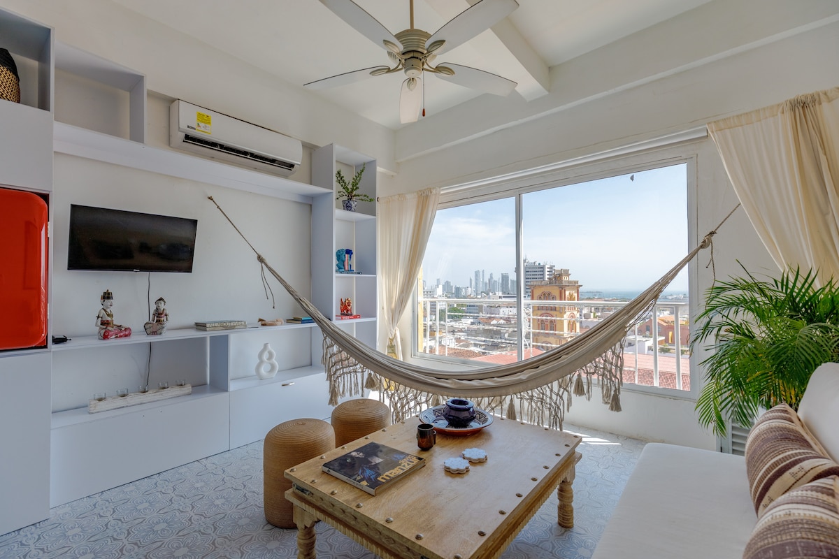 A spacious living area is highlighted by large windows offering expansive sea and city views. A light-colored sofa is positioned near a wooden coffee table with decorative items. A hammock is elegantly hung across the room, and a ceiling fan adds to the airy atmosphere.
