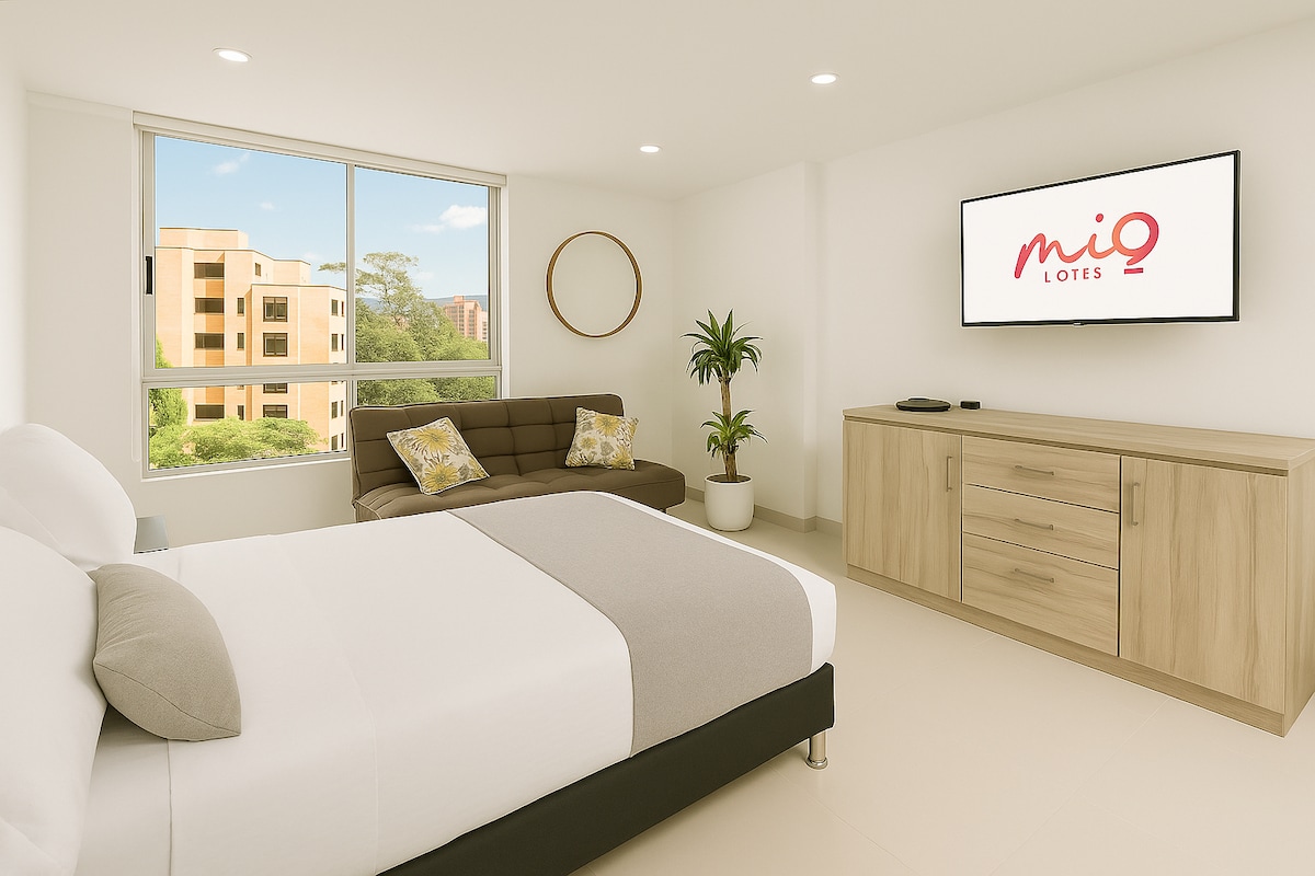 A bright sleeping area features a spacious bed with neutral bedding and light-colored pillows. A cozy sofa is positioned beside a large window that brings in natural light, with a wooden cabinet and a round mirror enhancing the simple decor. A television is mounted on the wall.